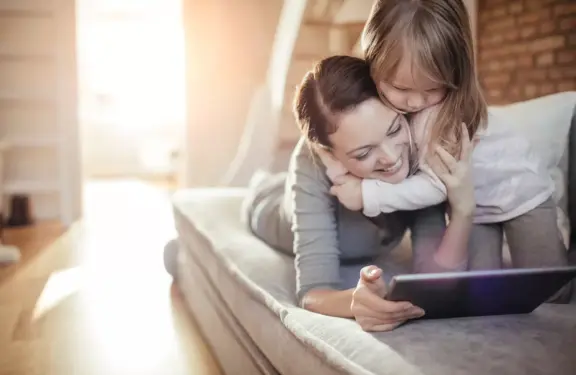 Dochter omhelst mama, terwijl ze samen op de bank zitten kijkend naar een tablet