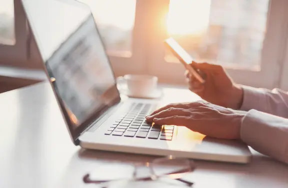 Man heeft linkerhand op laptop en in zijn rechterhand houdt hij zijn telefoon vast